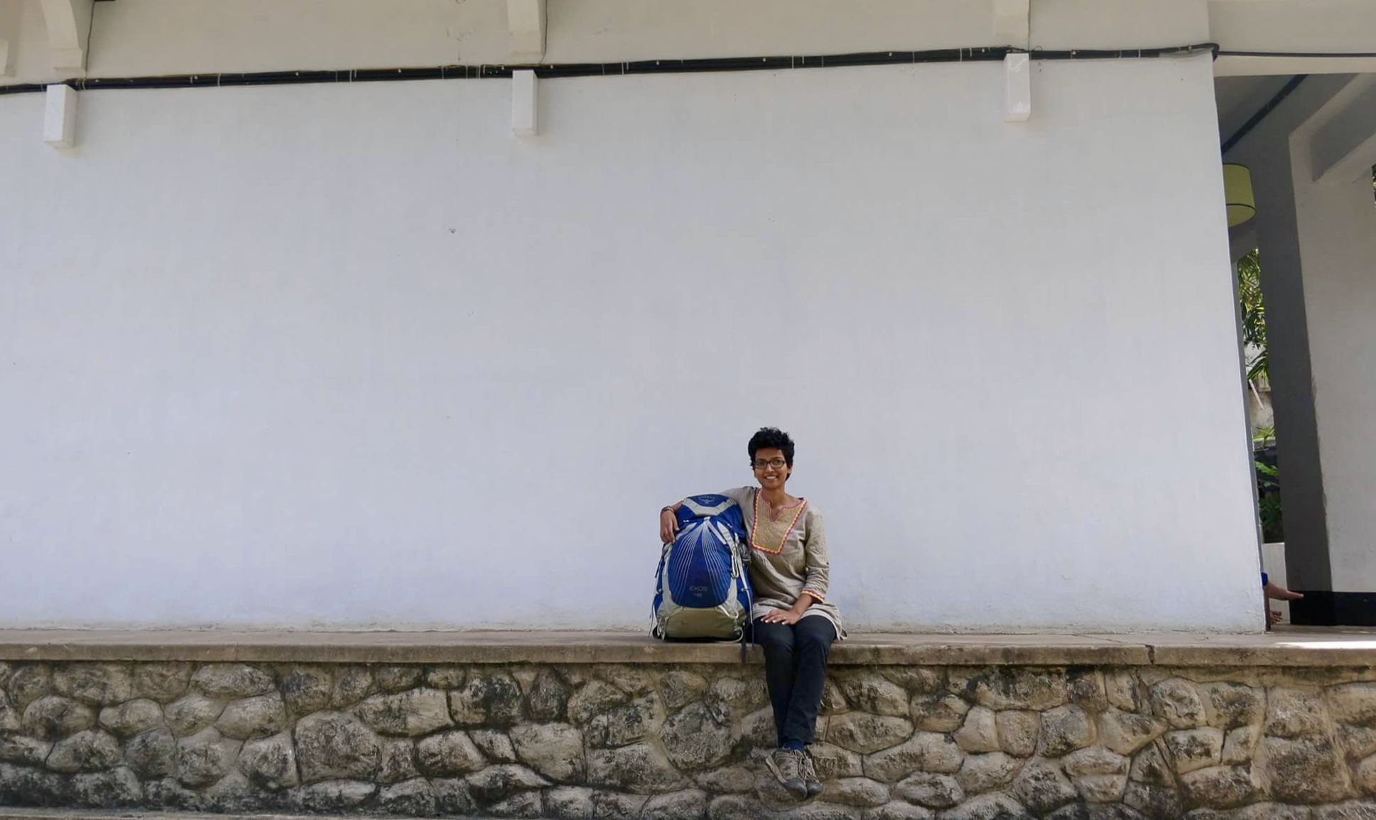 A woman with a blue backpack sitting on a stone platform against a white wall. 