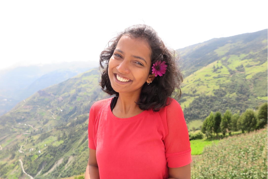 A woman smiling widely looking into the camera, a pink flower in her short hair and in the background is a green valley. 